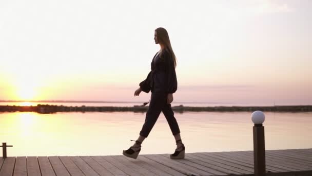 缓慢的动作, 镜头从侧面的美丽长发女子在黑色的木码头散步。清晨的黄昏, 在后台的阳光 — 图库视频影像