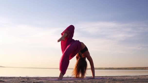 Close up view of a young woman in pink yogi pants practicing on sand near the sea or lake in the morning. Posture - Urdhva dhanurasana — Stock Video