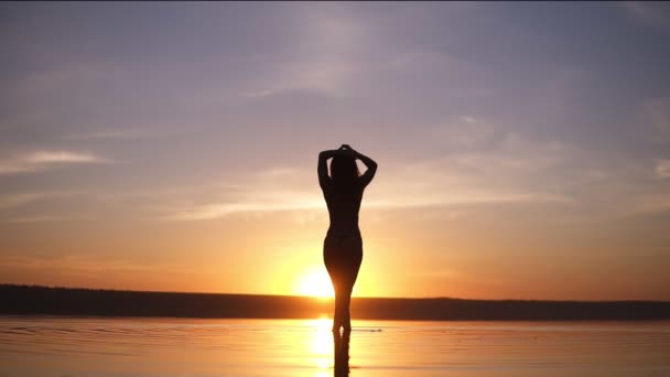 Silueta de una chica delgada contra una hermosa puesta de sol. Una chica se mete en el agua hasta el tobillo — Vídeos de Stock