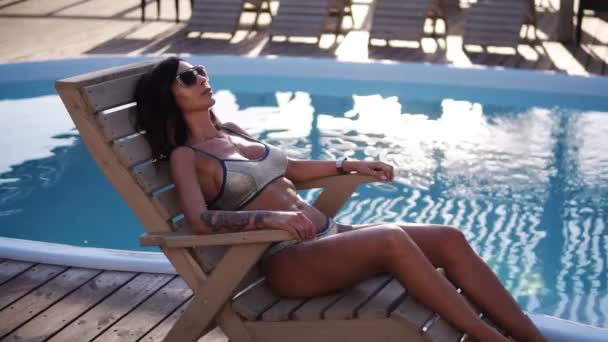 Havuzun yanında bir sehpa üzerinde çarpıcı, uygun bir beden ile genç bir kadın sunbathes. Bir kız bir şık moda mayo ve güneş gözlüğü. Su güneş parlar — Stok video