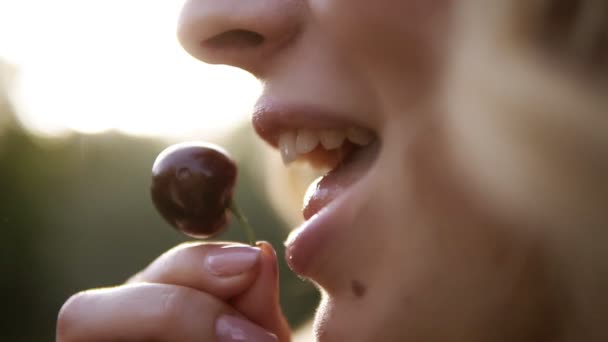 Extremadamente cerca de una joven rubia comiendo cereza. Mujer sexy sosteniendo una sabrosa cereza sobre un fondo de naturaleza borrosa. Al aire libre — Vídeos de Stock