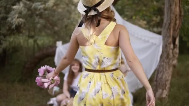Вечірка Хен у зеленому саду. Друзі дівчат зустрічаються зі своїм другом. Дівчина в жовтій сукні і літній капелюх щасливо приходить на пікнік з друзями — стокове відео