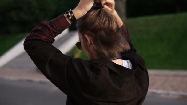 Grabación de una joven caminando al aire libre en la calle o en el parque. Alegre y feliz. Haciendo una cola de caballo en su pelo corto y claro, usando gafas de sol y ponerse los auriculares — Vídeos de Stock