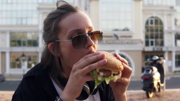 Молодая женщина ест большой, сочный гамбургер двумя руками. Молодая девушка в коротких волосах и темных солнечных очках. Голоден. Закрыть — стоковое видео