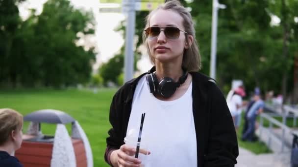 Vorderseite Tracking-Aufnahmen von einem jungen, hübschen Mädchen, das durch den Park geht. mit Kopfhörern am Hals und stylischer Sonnenbrille. Freizeit — Stockvideo