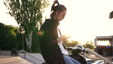 Bir motosiklet üzerinde oturan ve onun cep telefonuna yazarak siyah kapüşonlu genç kız. Motosiklet parkta oturan. Güneş ışınları ve arka plan üzerinde