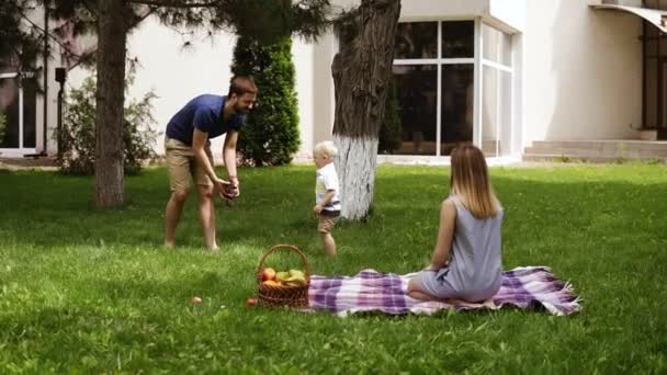 Vista frontal de una familia idilio pasando el rato juntos en el parque. Joven mamá sentada en una hierba y mirando a su hijo jugar. El padre divierte al hijo, lanza conos al aire. Movimiento lento — Vídeo de stock