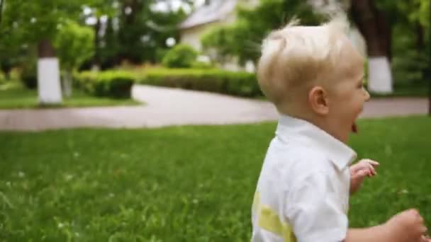 Fröhlicher, lächelnder blonder Junge, der fröhlich mit seiner zur Mutter hängenden Zunge rennt. Glücksmomente. Mutter umarmt und küsst ihr Kind. Grüner Park — Stockvideo