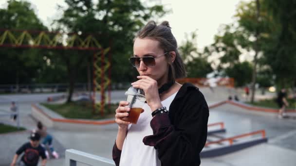 Een stijlvolle meisje in zonnebrillen en hoofdtelefoon geniet van een drankje in een transparant glas, dranken met een rietje. Een vrouw staat in het midden van een park met schaatsers en kinderen op skateboards. Permanent — Stockvideo