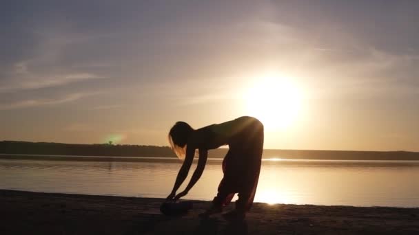 Mujer joven haciendo ejercicio de yoga - abriendo su esterilla de yoga cerca del mar o el océano. Puesta o salida del sol. Concepto de salud. Movimiento lento . — Vídeos de Stock