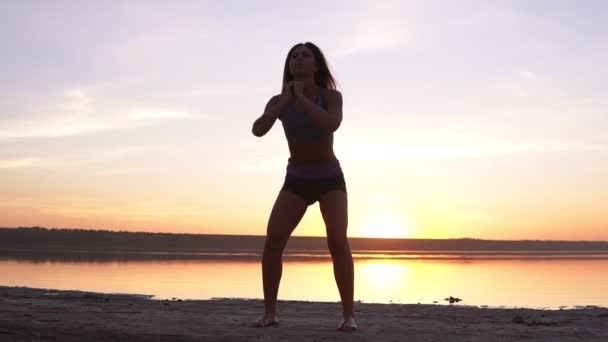 Giovane donna attraente che fa esercizi sportivi al mattino sulla spiaggia all'alba sul mare o sul lago. Facendo sit up in abbigliamento sportivo. Stile di vita sano, vita attiva, esercizio fisico — Video Stock