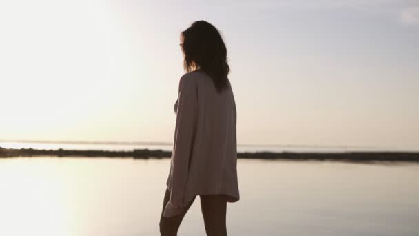 清晨的薄雾中, 一个女孩穿着浅色的长袍, 沿着海边的泳装散步。美丽的黑发在早晨黄昏和阳光摆在游泳衣 — 图库视频影像