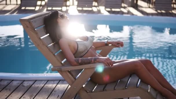 Filmati di moda di una bella donna abbronzata dai capelli castani in elegante bikini argentato che si rilassa accanto a una piscina con occhiali da sole. Il sole splende e lampeggia su un'acqua — Video Stock