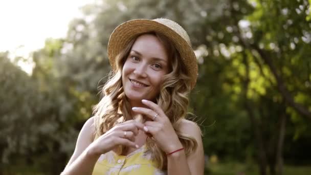 Hermosa mujer rubia sonriendo en verano soleado o día de primavera afuera en un prado. Chica con sombrero de paja en la naturaleza. Posando en cámara, sonriendo. Fondo de árboles verdes. Vista frontal — Vídeos de Stock