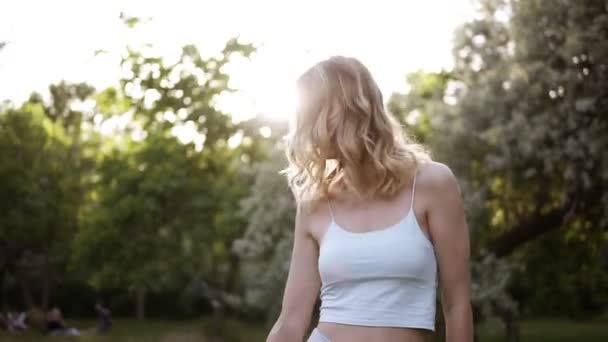 Озил, блондинка в короткой белой рубашке, счастливо гуляющая по зеленому парку или лесу. Подбрасывает небольшой букет цветов в воздух. Медленное движение — стоковое видео