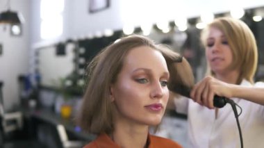 İle çekici beyaz kız makyaj güzellik salonunda. Kuaför saç demirle düzeltiyor. Arka plan bulanık - beyaz lambalar ve aynalar