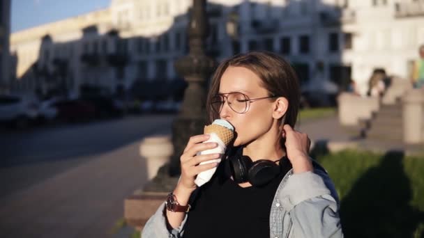 Giovane donna in camicia arancione godendo morbido gelato alla vaniglia in cono waffle all'aperto al rallentatore. Donna che mangia un gelato in strada da sola, seduta su una panchina in città — Video Stock