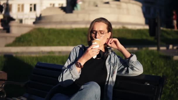 Mujer joven en ropa casual disfrutando de su tiempo libre, sentada en un banco de la calle y lamiendo helado de vainilla. Delicioso postre. Día soleado — Vídeo de stock