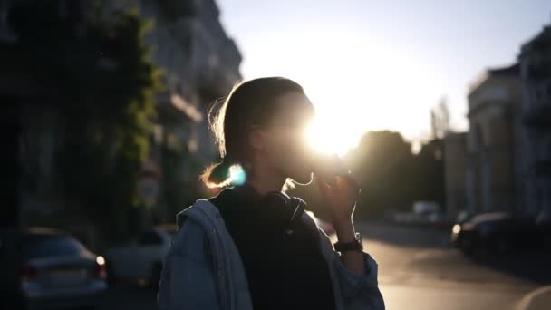 Een meisje in stijlvolle glazen eet ijs in een kegel wafel in de stralen van de zonsondergang. Casual kleding. Wandelen in de stad. Zijaanzicht — Stockvideo