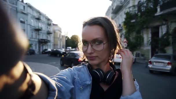 Una joven con gafas posa con una cámara en su mano extendida. Sonriendo, feliz, muestra un gesto de mano "Cool". Atardecer — Vídeos de Stock