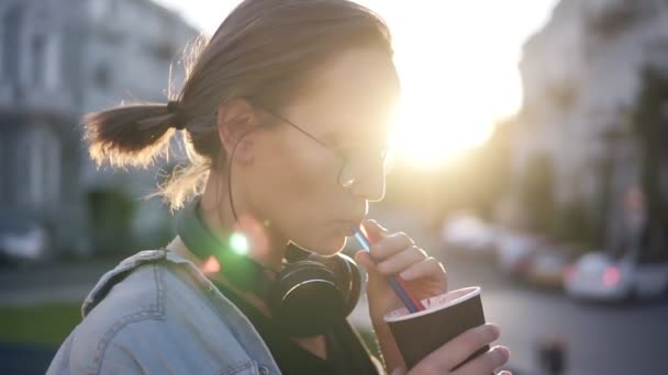 Vue latérale d'une belle jeune femme avec un casque sur le cou en train de boire une boisson à l'aide d'une paille. Une blonde en ville. Soleil brille sur le fond — Video