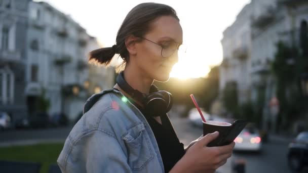 Genç ve güzel kız mavi gömlek ve kulaklık şehrin sokak üzerinde duran ve cep telefonu kaydırma boynunda. Bir kahve elinde tutuyor. Yan görünümü. Dusk — Stok video