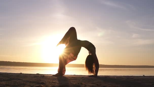 junge Frau in schönen farbigen Yogahosen macht Yoga-Übung urdhva dhanurasana, auch bekannt als Bogenhaltung nach oben an einem leeren Strand. Sonnenlicht auf dem Hintergrund