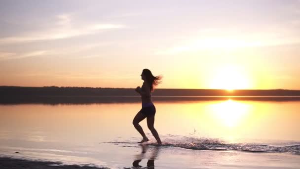 Mujer delgada con el pelo largo corriendo al aire libre. Correr junto al agua. Junto al mar. El sol brilla en el fondo — Vídeos de Stock