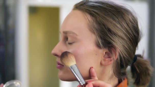 Vista lateral de una joven y bonita modelo femenina. Proceso de maquillaje. Artista poniendo un poco de colorete transparente y brillante en la mejilla de las mujeres usando un pincel — Vídeos de Stock