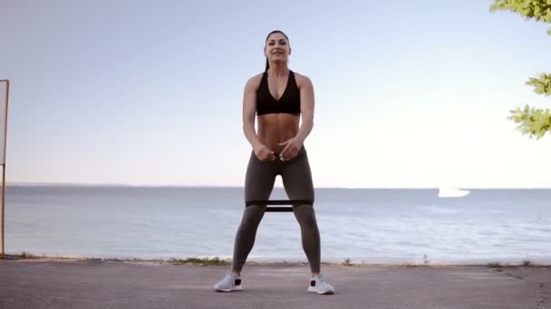 Hermosa mujer musculosa disfrutando de su ejercicio al aire libre cerca del mar. Hacer sentadillas con ropa deportiva, entrenar sus piernas y sonreír mientras hace sentadillas con goma. Mar u océano en el fondo — Vídeo de stock