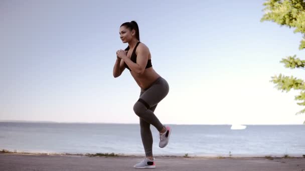 Beautiful fitness girl doing exercises on a seashore. Full length of a professional sports woman stretching her legs with a rubber. Summer, bright day — Stock Video