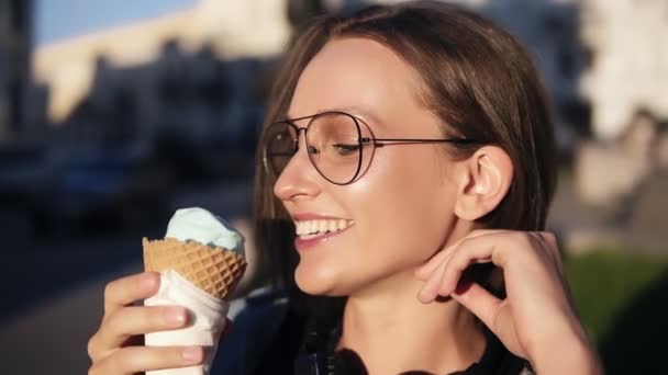 Запись, на которой привлекательная девушка ест большое белое мороженое на улице. Молодая женщина в современных прозрачных очках, улыбающаяся, наслаждающаяся своим временем — стоковое видео