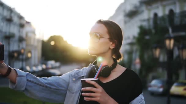 Porträt einer jungen attraktiven Frau mit Sonnenbrille und Kopfhörern auf dem Hals, die in der Stadtstraße vor der Kamera posiert. Nahaufnahme — Stockvideo