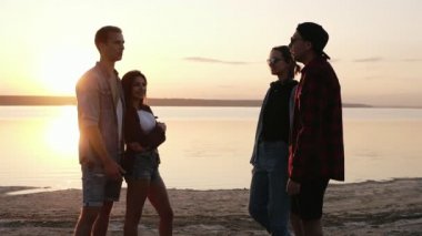 Grup arkadaşıyla birlikte sahilde asılı. Su üstünde gün batımı. Güzel iki çift. Konuşmuyor, el hareketi. Dostluk kavramı