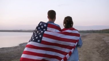 Deniz kıyısında yürüyüş, el ele tutuşarak, mesafe içine bakarak bir çift. Arkadan ateş. Genç çift Amerikan bayrağı ile kaplı