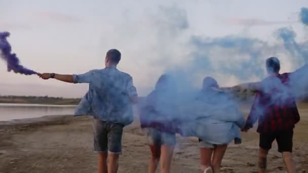 Quattro amici in abiti casual divertirsi insieme. Correre e tenersi per mano. I ragazzi tengono tra le mani dei bracci di fumo blu. Vista posteriore — Video Stock