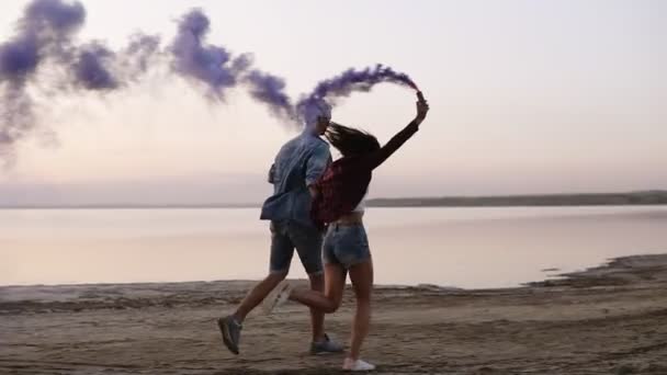 Giovane coppia felice in esecuzione con granata di fumo colorata nelle loro mani in riva al mare durante il tramonto. Divertente, fumo colorato intorno a loro — Video Stock