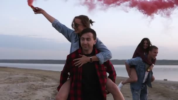 Belos jovens casais com bombas de fumaça nas mãos na praia. Amigos - homens e mulheres estão brincando com fumaça colorida, piggyback. Emoções, estilo de vida feliz — Vídeo de Stock