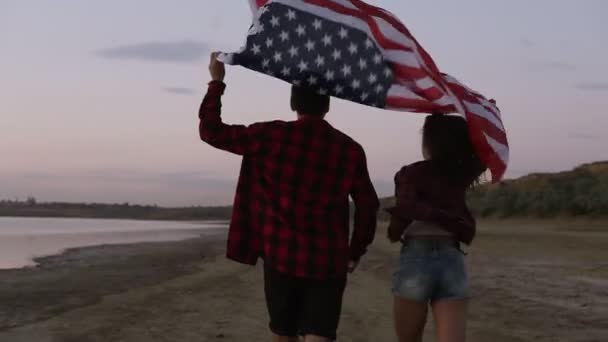 Jong koppel beide in rode geruite overhemden lopen aan de kust met Amerikaanse vlag boven hun hoofd. Weergave achterzijde — Stockvideo