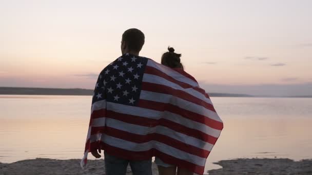 Närbild av vackra unga paret ser fram emot och drömmer. Omfamnade älskare funderar på deras framtida liv stående vid havet i lugna kvällen, täckt med amerikanska flaggan — Stockvideo