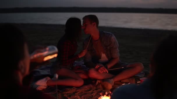 Bonita pareja caucásica alimentándose mutuamente con malvavisco en palo, besándose frente a una hoguera. Amigos están pasando el rato juntos en la playa. Atardecer — Vídeos de Stock