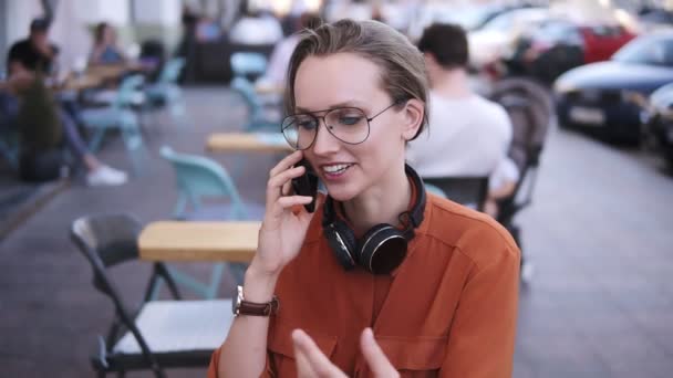 Женщина со смартфона разговаривает по телефону, сидя в кафе. Она улыбается. Красивая молодая женщина, разговаривающая по мобильному телефону. Толпа на улице, на улице — стоковое видео