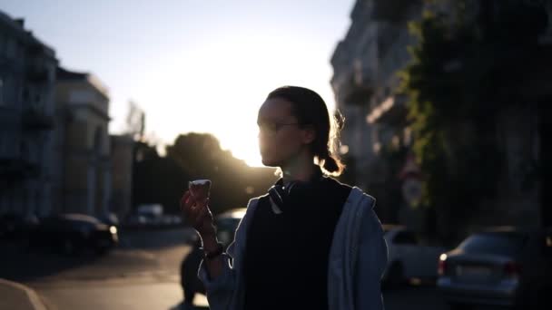 Современная, стильная девушка, стоящая на улице под солнечным светом и кушающая мороженое. Тени солнца на зданиях, закат. Медленное движение — стоковое видео