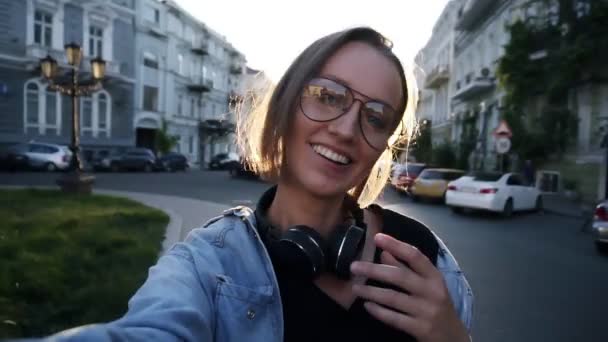 Retrato de feliz atraente jovem posando para câmera. Um estilo de vida sorridente e feliz. Mulher bonita sorrindo para a câmera com fones de ouvido no pescoço, vestindo óculos modernos e transparentes — Vídeo de Stock