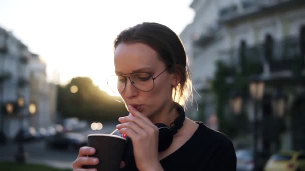 Jonge vrouw in stijlvolle zonnebril drinken van een kopje koffie met plastic rietjes op de straat. Avondschemering. Zon schijnt op de achtergrond — Stockvideo