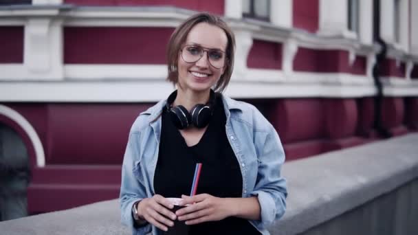 Vista deslumbrante de uma jovem menina bonita em óculos de pé com um vermelho, edifício clássico no fundo e sorrindo para a câmera. Roupas casuais, fones de ouvido no pescoço, segurando uma xícara com uma palha — Vídeo de Stock