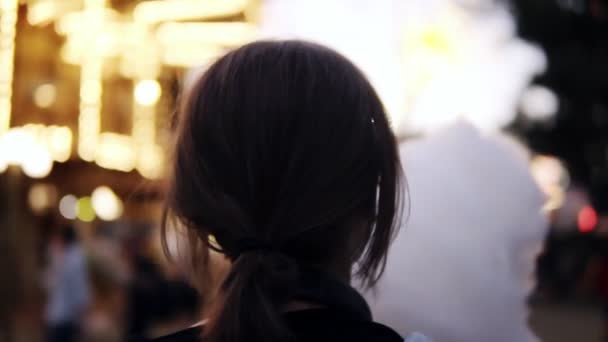 Vista trasera de una chica caminando en el parque de atracciones, comiendo un algodón de azúcar. Mujer rubia de pelo corto con gafas transparentes — Vídeos de Stock