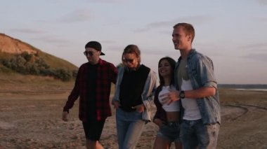 Bir grup genç insan deniz veya göl yakınında kumlu karayoluyla yürüme. Gülümseyen iletişim. Genç bir çift. Rahat kıyafetler, gençlik kültürü giyiyor