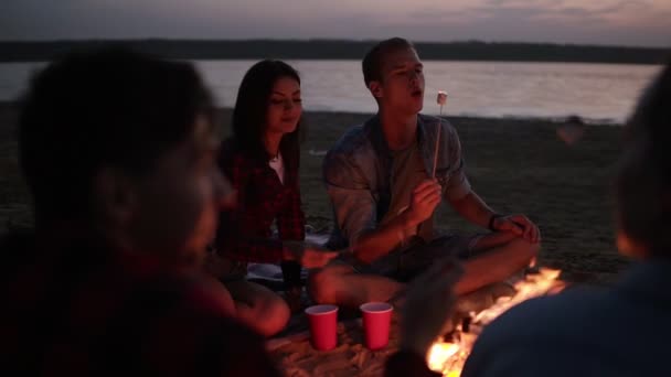Mooie paar op een vreugdevuur, marshmallows roosteren zoenen. Jongeren zitten in een kring van vrienden op het strand, 's avonds tijd — Stockvideo