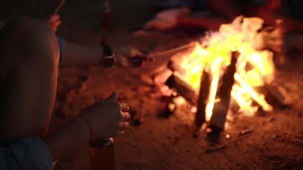 Close up of a young hipster female with headphones on her neck frying marshmallow on stick from bonfire. Friends are sitting near the fire, picnic concept. Night time — Stock Video